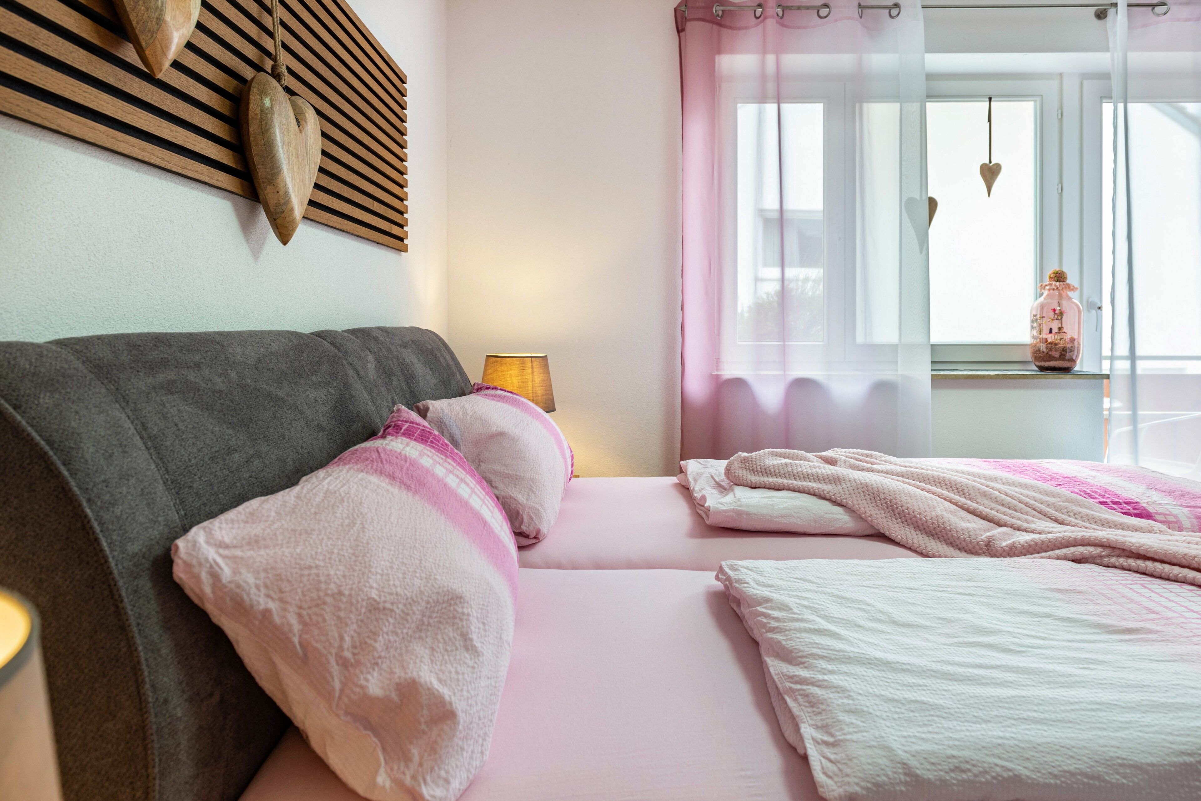 A bedroom with a bed, pillows, a bedside lamp, a window with curtains, and a wall-mounted wooden panel.