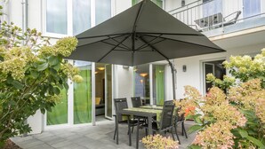 An outdoor patio with a table and chairs, a large umbrella, and greenery. - Apartment 'Jasmin' with Large Terrace and View of the Beautiful Flower Garden (Sipplingen am Bodensee)