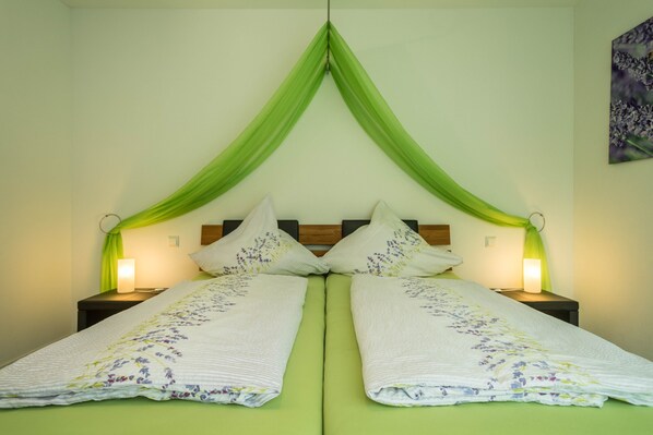 A double bed with green curtains, two bedside lamps, and a floral bedspread. - Cozy, Lovingly Furnished Holiday Apartment "Lavendel" with Covered Terrace & Balcony (Sipplingen am Bodensee)