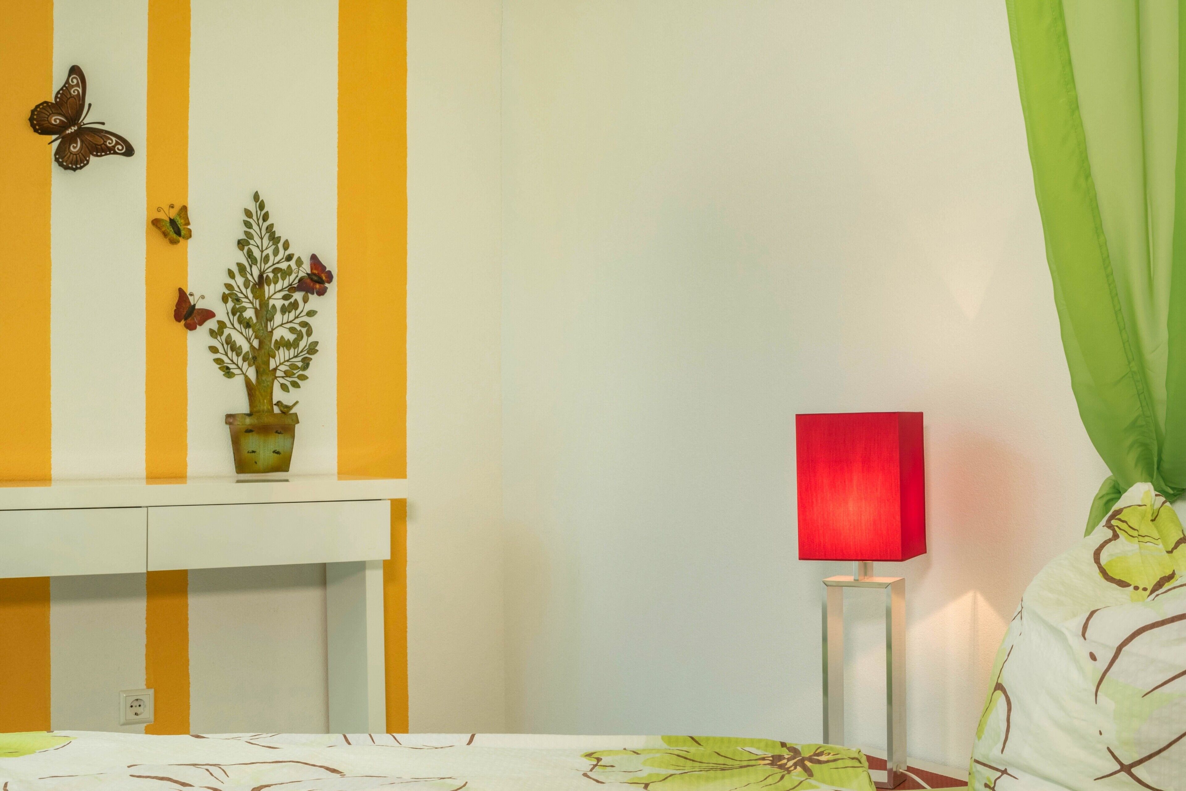 A room with a bed, a bedside table with a lamp, a wall with vertical stripes, and a potted plant.