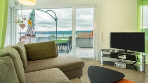 Una sala de estar con un sofá, un televisor sobre un soporte de vidrio y vistas a un patio con una mesa y sillas.