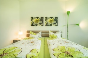 Une chambre à coucher avec un lit, deux tableaux représentant des fleurs accrochés au mur, une lampe de chevet et une grande plante verte.