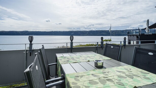 An outdoor terrace with a table and chairs, overlooking a body of water and a distant shoreline. - Apartment "Rose" with Large Balcony and Stunning Lake, Forest, and Mountain Views (Sipplingen am Bodensee)