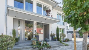 A modern building with a glass entrance, outdoor plants, and a visible number '6' on the facade. - Apartment "Rose" with Large Balcony and Stunning Lake, Forest, and Mountain Views (Sipplingen am Bodensee)