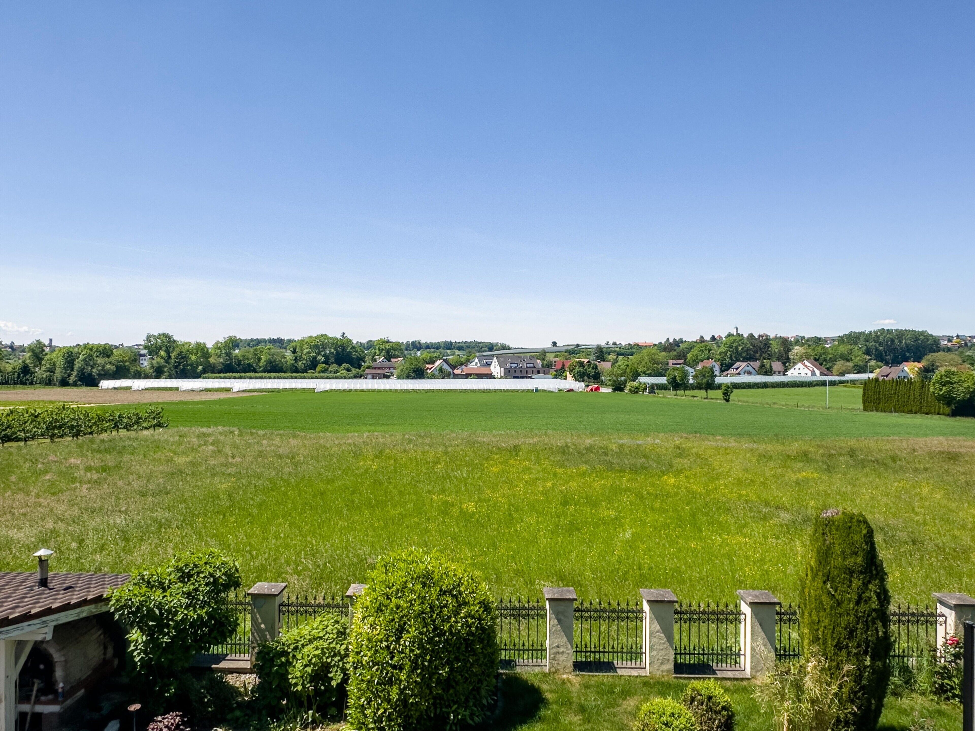 A large green field with a white building in the distance, surrounded by trees and a clear sky.
