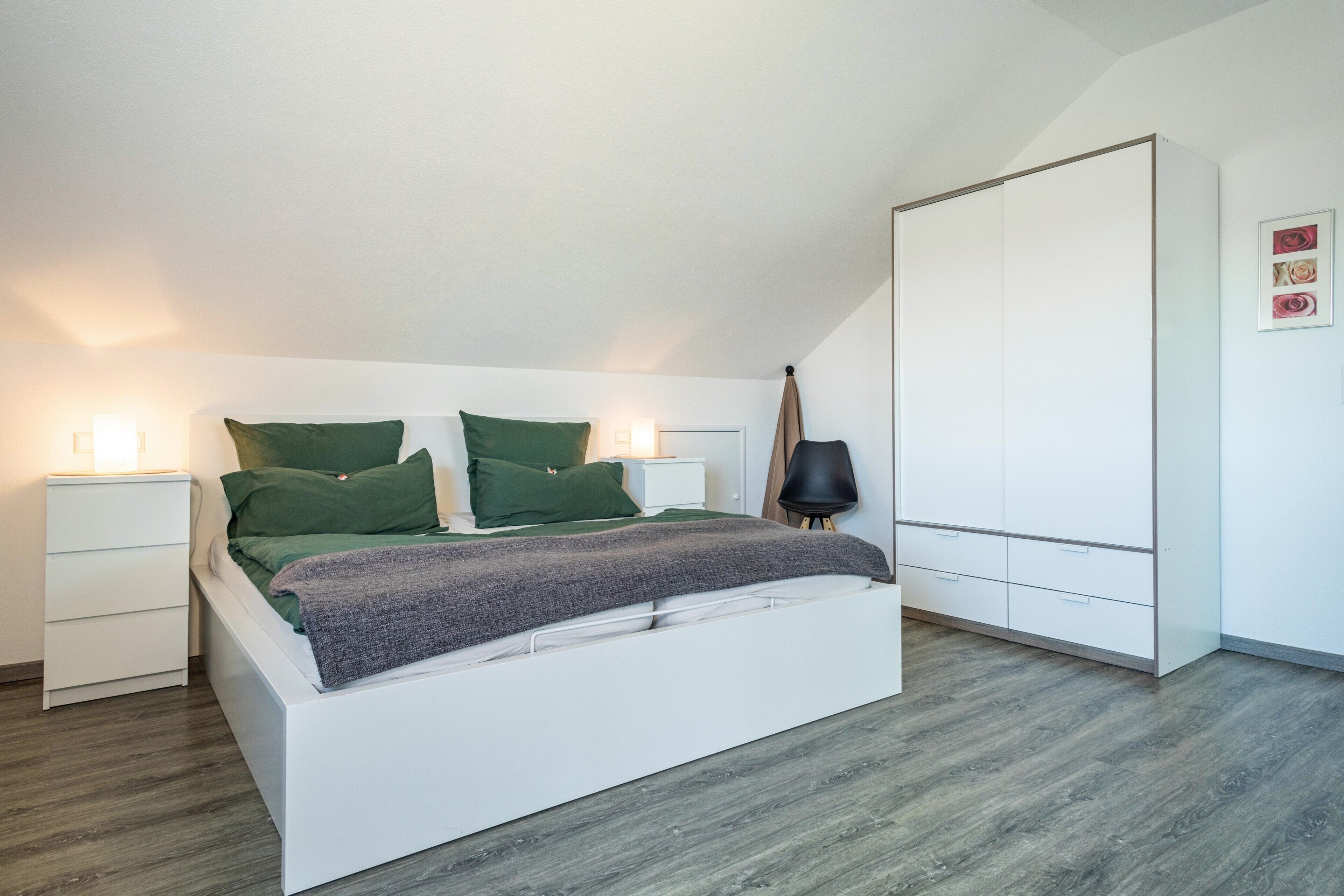 Ein Schlafzimmer mit einem weißen Bett, grünen Kissen, einem Nachttisch, einer Lampe und einem Kleiderschrank.