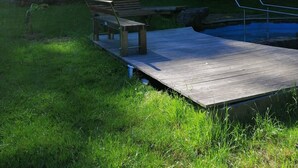 En træbænk og en træplatform ved vandet, omgivet af græs og træer.