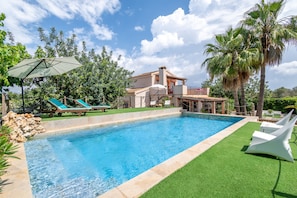 A swimming pool with lounge chairs, a covered area with an umbrella, and a house with a balcony.