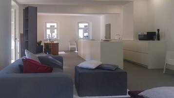 A modern living room with a grey sofa, ottoman, and a small table with a lamp. There is a kitchen area with a sink and a countertop.