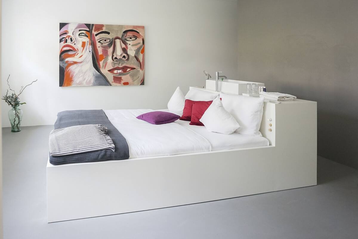 Ein modernes Schlafzimmer mit einem weißen Bett, vielen Kissen, einem Nachttisch und einem an der Wand hängenden Kunstwerk.