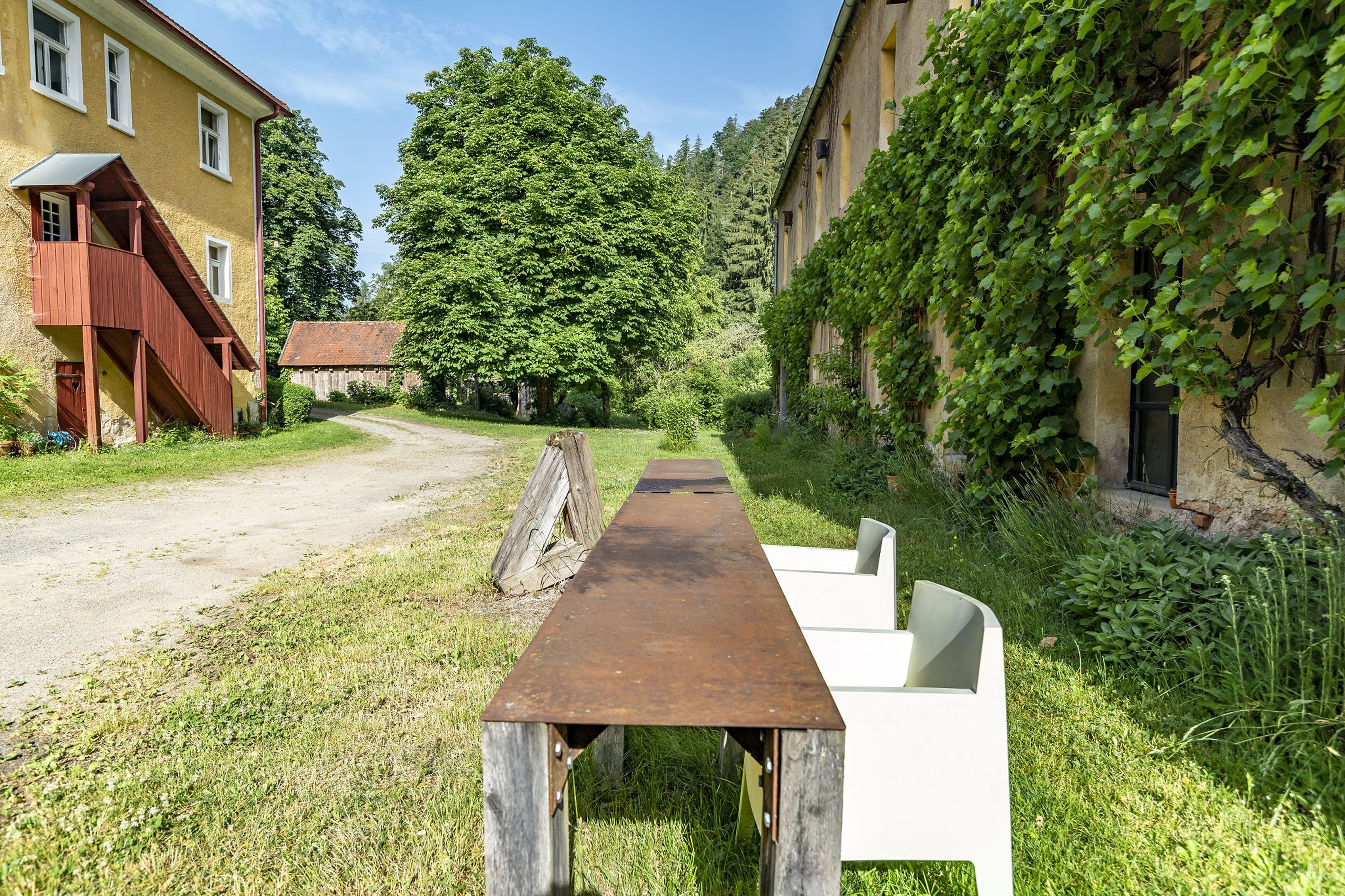 Un cadre extérieur rustique, avec un banc en bois et une chaise blanche, entouré de verdure et d’un bâtiment doté d’un escalier.
