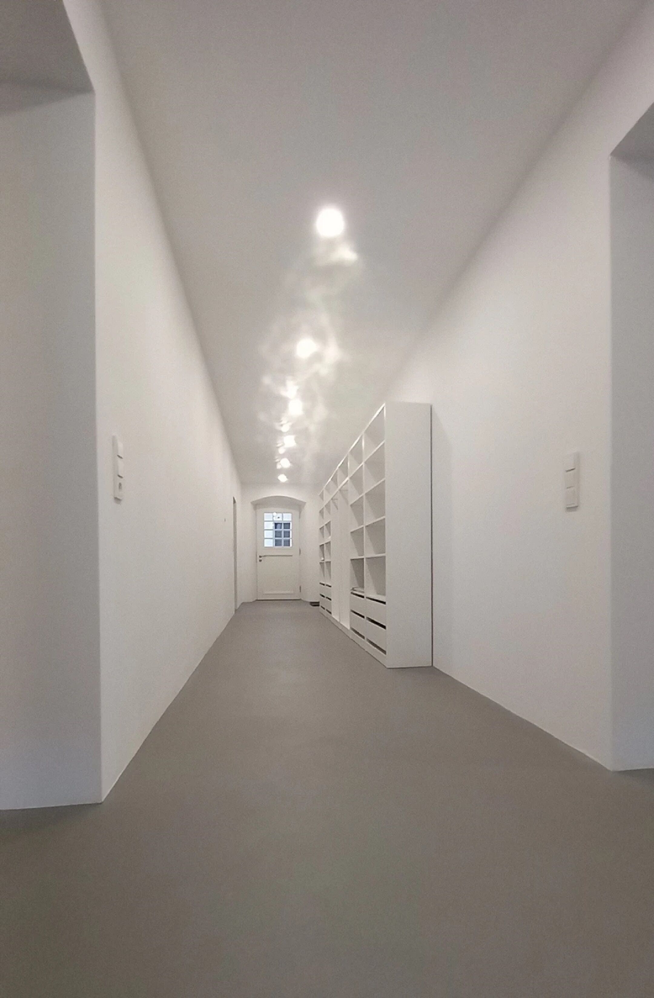 Un pasillo estrecho con paredes blancas y una lámpara en el techo. Hay una puerta al final y una ventana a la izquierda.