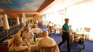 A dining area with tables and chairs, a person walking, and a wheelchair. - Residenz Am Malerwinkel (Bad Sassendorf)