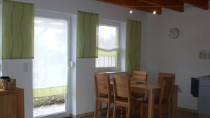 A dining area with a table and chairs, a window with curtains, a clock on the wall, and a small kitchenette. - Ferienanlage Karolinenhof 2 (Haundorf)
