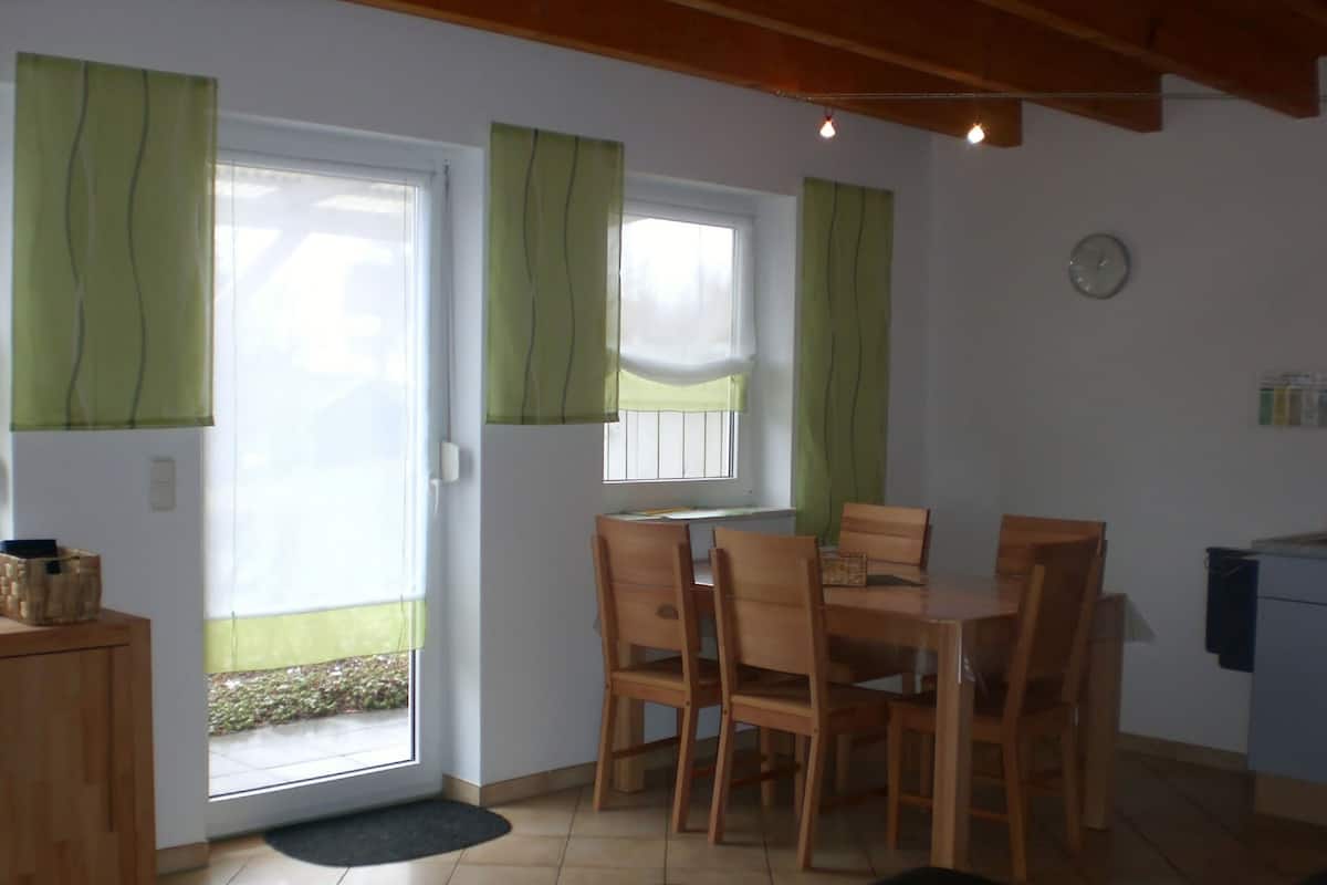A dining area with a table and chairs, a window with curtains, a clock on the wall, and a small kitchenette.