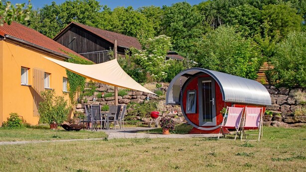 A small, red, cylindrical building with a door and windows, situated in a grassy area with a yellow building and a wooden structure in the background. - Pferdefreunde Loberhof (Weihenzell)