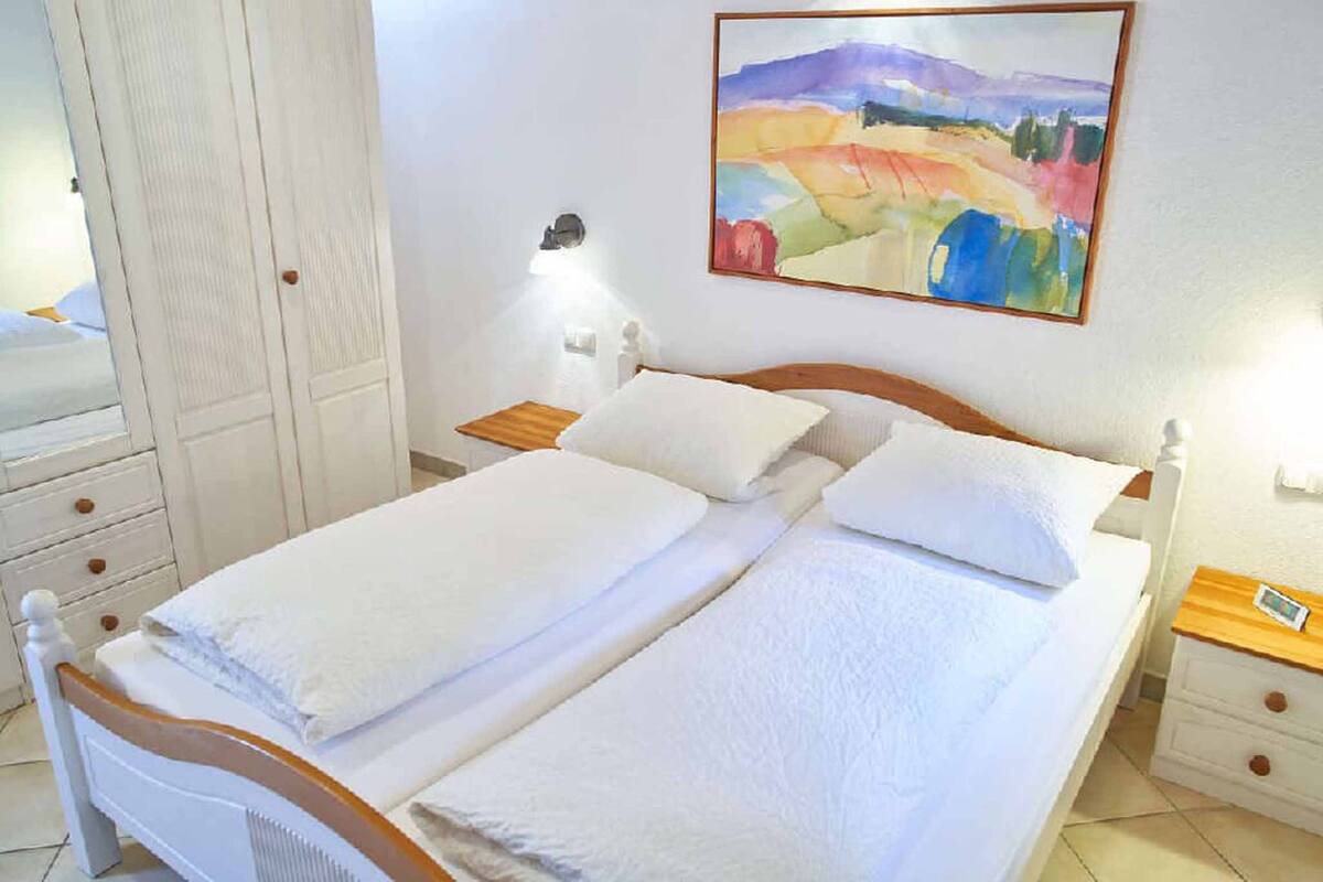 Una cama bien hecha con sábanas blancas, una mesita de noche con un reloj y un cuadro colgado en la pared.