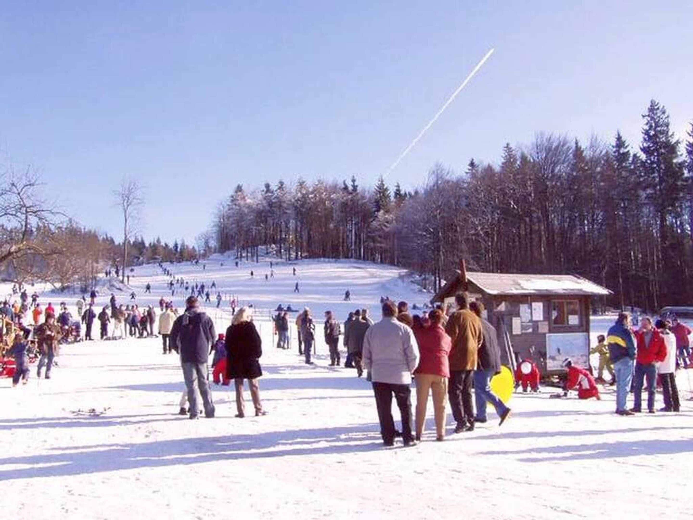 A snowy outdoor area with a small wooden structure, likely a lodge or information center, surrounded by people and a ski slope in the background.