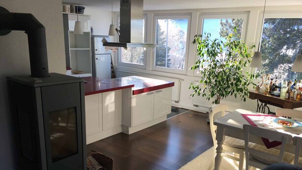 Une cuisine moderne avec des armoires blanches, un plan de travail rouge et une cuisinière intégrée. On y trouve un coin repas avec une table et des chaises, ainsi qu’une grande fenêtre qui laisse entrer la lumière naturelle.