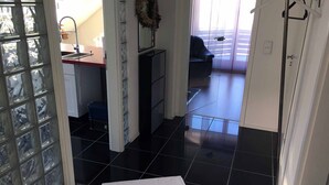 Une pièce avec un sol carrelé, une table blanche, un coin cuisine avec un évier et une vue sur l’extérieur, visible à travers une porte vitrée.