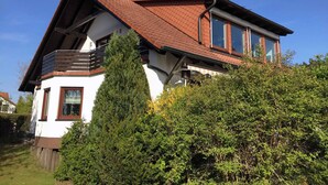 Une maison à deux étages, dotée d’un toit en pente, d’un balcon à l’étage supérieur et d’une haie verdoyante et dense au premier plan.