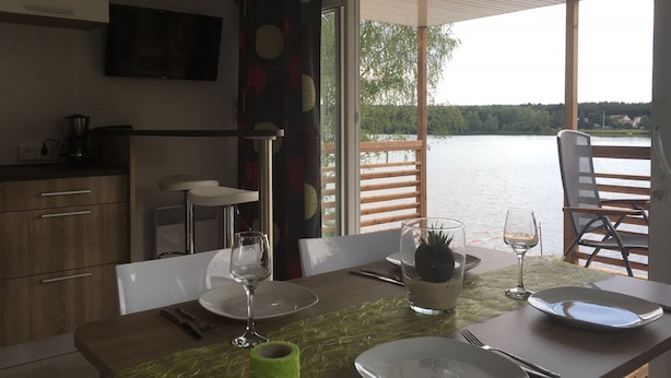 A dining area with a table set for two, a view of a lake, and a kitchen area with a coffee maker. - Pension Lutter (Bodenwoehr)
