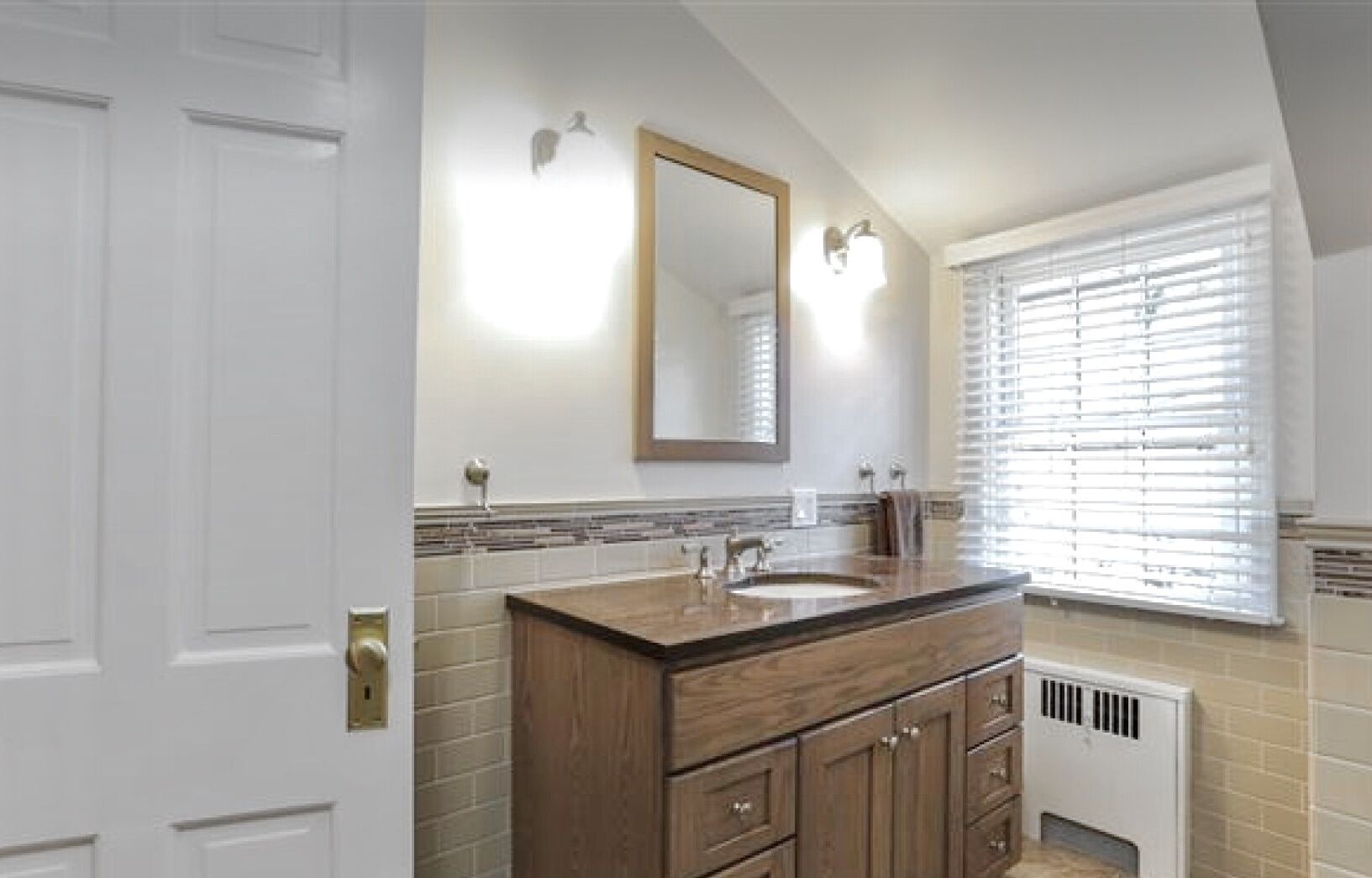 A bathroom with a wooden vanity, a large mirror, and a window with blinds.