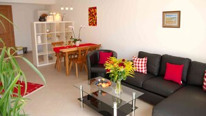 Un salon moderne avec une table basse en verre, un canapé noir agrémenté de coussins rouges, un coin repas avec des chaises en bois et une étagère blanche.