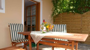 Une table en bois recouverte d’une nappe blanche, deux chaises à rayures et un banc. Une bouteille de vin et un vase rempli de fleurs sont posés sur la table.