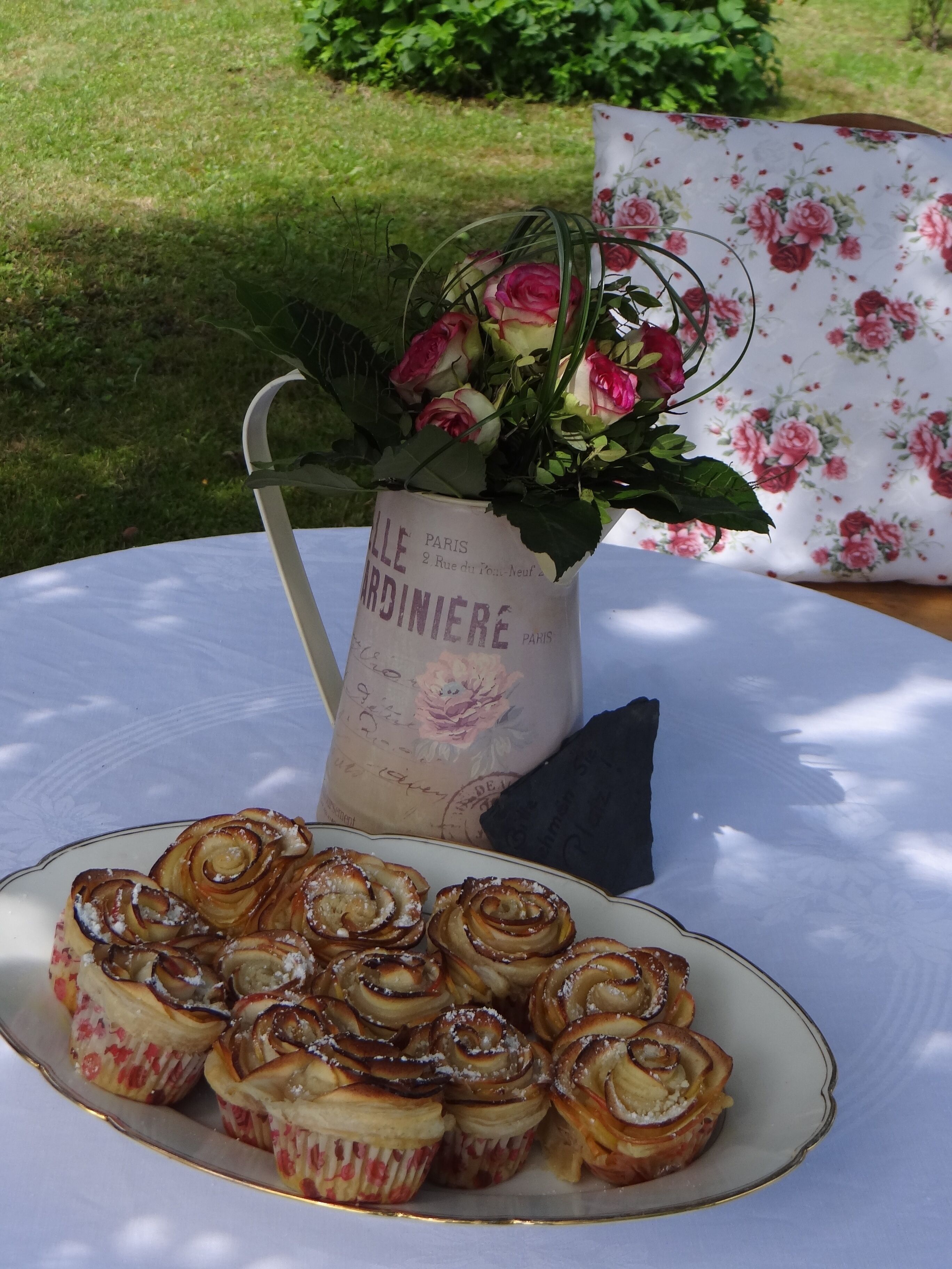 Una mesa con mantel estampado de flores, un florero con rosas rosadas y un plato de rollos de canela.