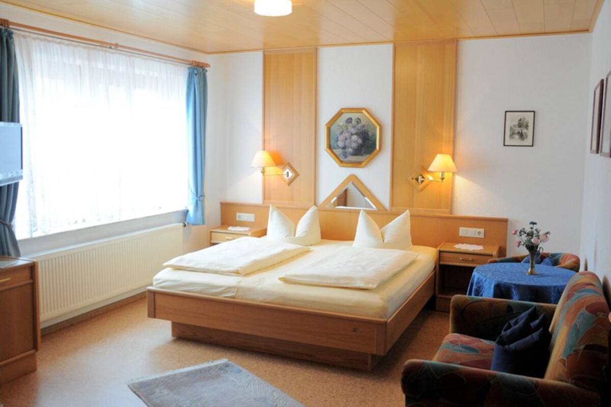 Una habitación de hotel con una cama grande, dos mesitas de noche, una pequeña mesa con un florero, una silla y una ventana con cortinas.