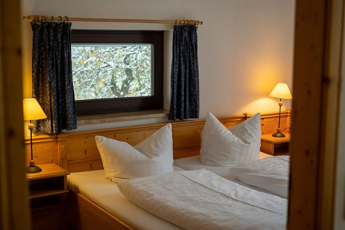 Un dormitorio con cabecera de madera, dos mesitas de noche con lámparas, una ventana con cortinas y una cama con almohadas.