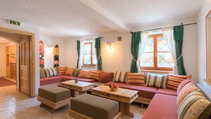 A cozy room with a red sofa, a wooden bench, and a small table with a fruit bowl. There are two windows with green curtains, and a wooden door leading to another room. - Chalet Murr by Skinetworks (Sankt Anton am Arlberg)