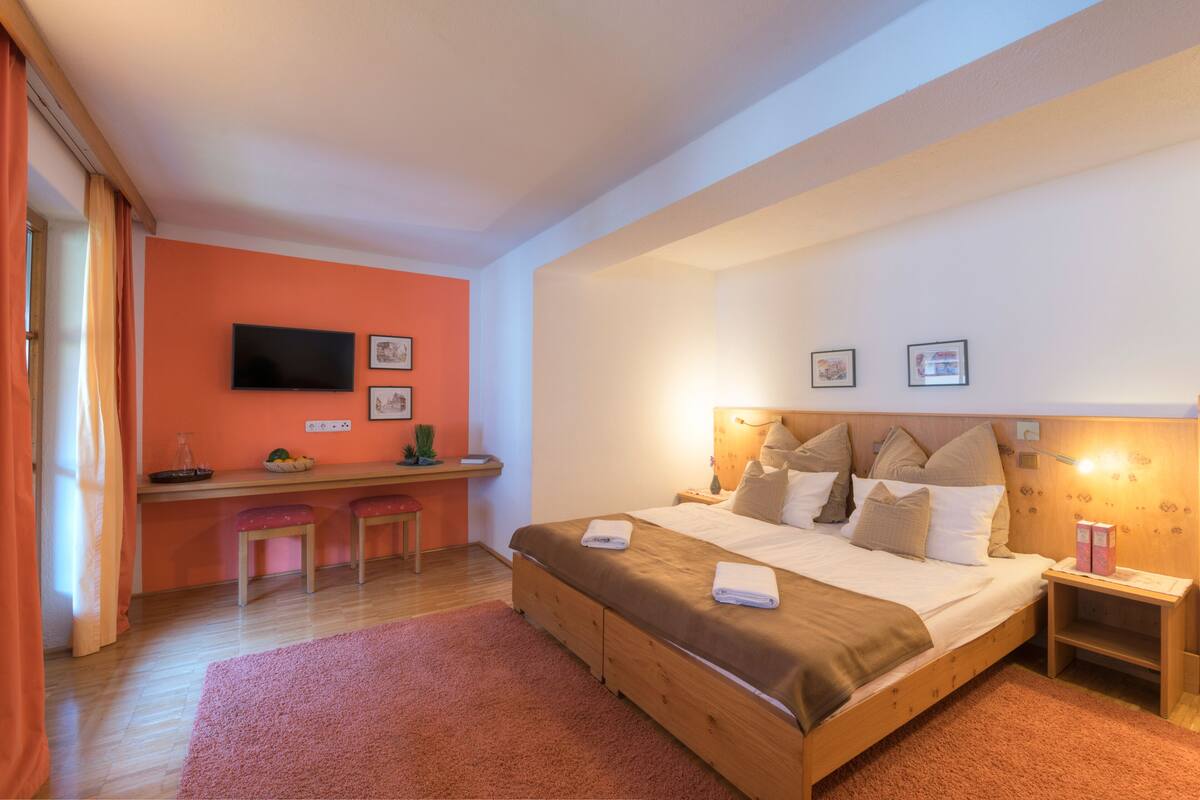 Un dormitorio con cama, mesitas de noche, un escritorio con sillas, televisión y obras de arte en las paredes.