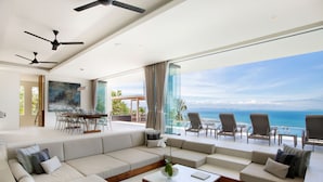 A modern living room with a large sectional sofa, a coffee table, and a dining area with chairs. There are ceiling fans and a large window with a view of the ocean. - Villa Zest at Lime Samui (Koh Samui)
