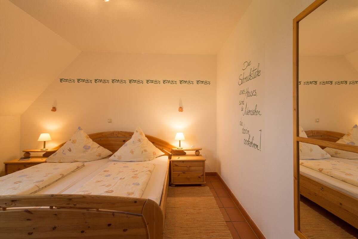 A bedroom with two wooden beds, each with a nightstand and a lamp. The room has a sloped ceiling and a mirror on the wall.