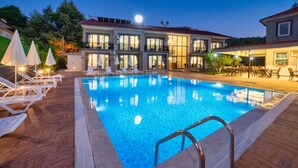 Une piscine entourée de chaises longues et de parasols, elle-même bordée d’un bâtiment doté de grandes fenêtres et d’un espace de détente extérieur.