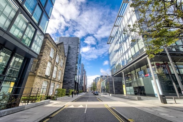 A modern urban street with buildings, a clear sky, and a road with double yellow lines.