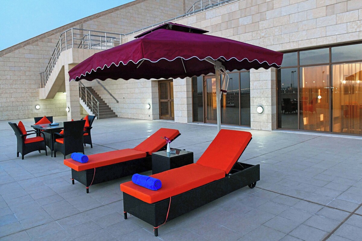 Un área de estar al aire libre con sillones reclinables y mesas, una escalera y la fachada de un edificio al fondo.