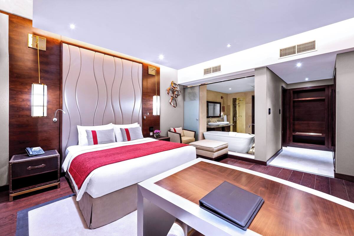 A modern hotel room with a large bed, bedside table, and a desk with a laptop. There is a bathroom visible through an open door.