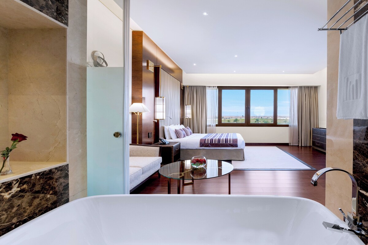 Una habitación de hotel moderna con una cama grande, una zona de estar con una mesa de vidrio, una bañera y vistas al exterior.
