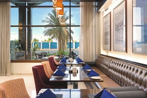 A dining area with a long table set for a meal, cushioned seating, and large windows offering a view of palm trees and a blue building.