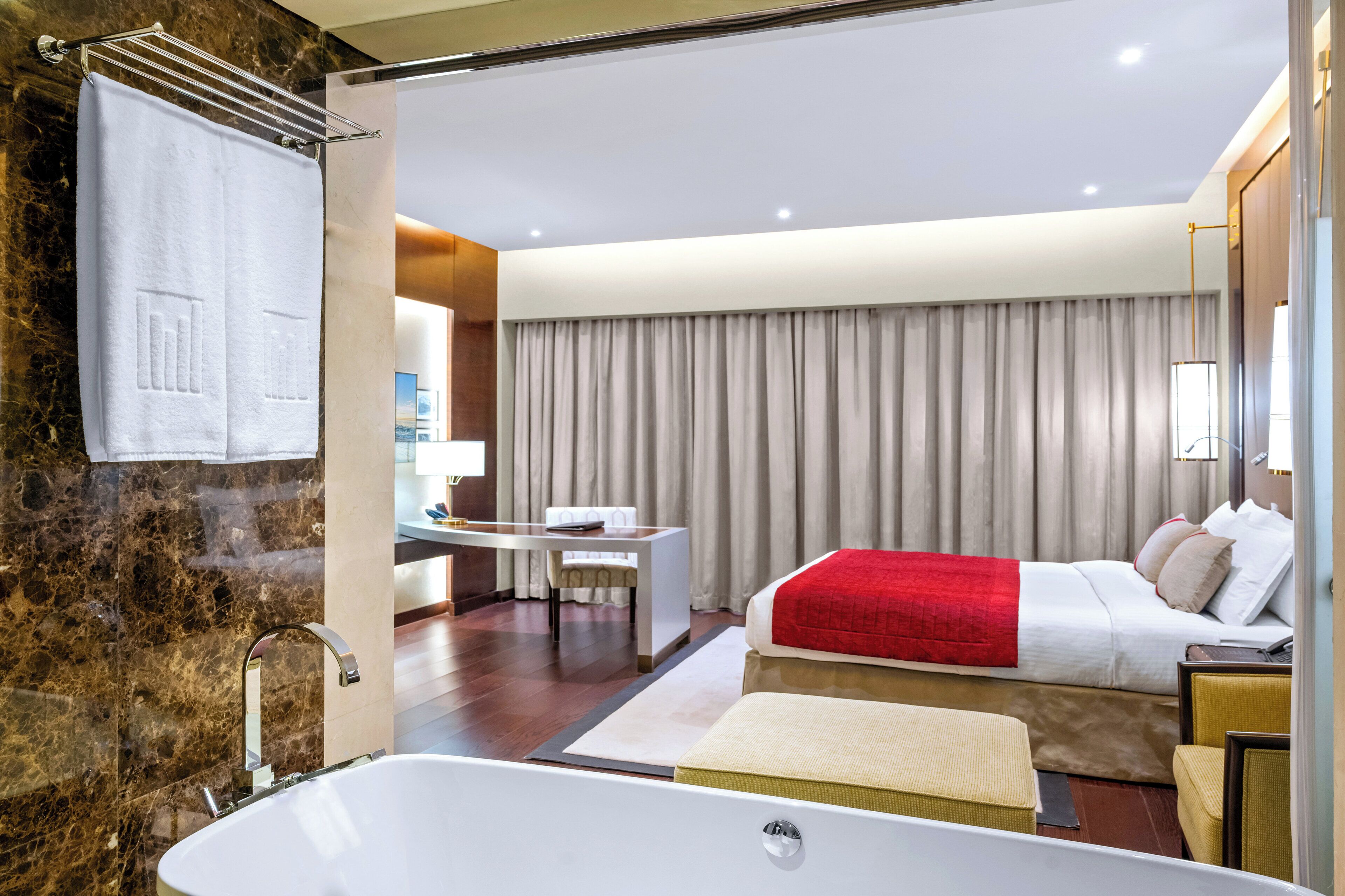 Ein modernes Hotelzimmer mit Marmorbad, einem Bett mit roter Decke, einem Schreibtisch und einem großen Fenster mit Vorhängen.