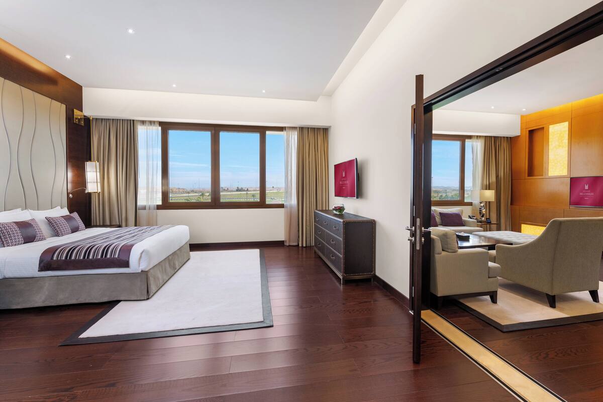 A modern hotel room with a large bed, a sitting area with a sofa and armchair, a dresser, and a large mirror reflecting the room.