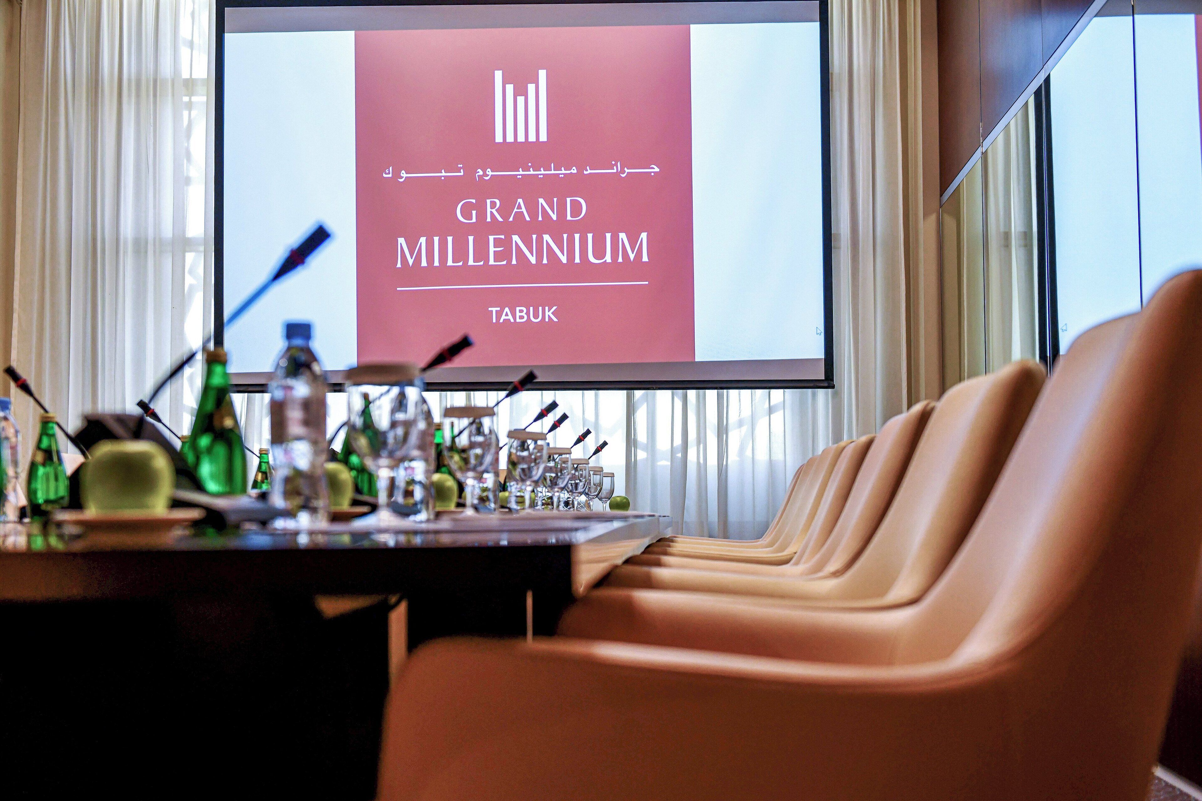 A conference room with a projection screen displaying "GRAND MILLENNIUM TABUK" and a row of leather chairs.