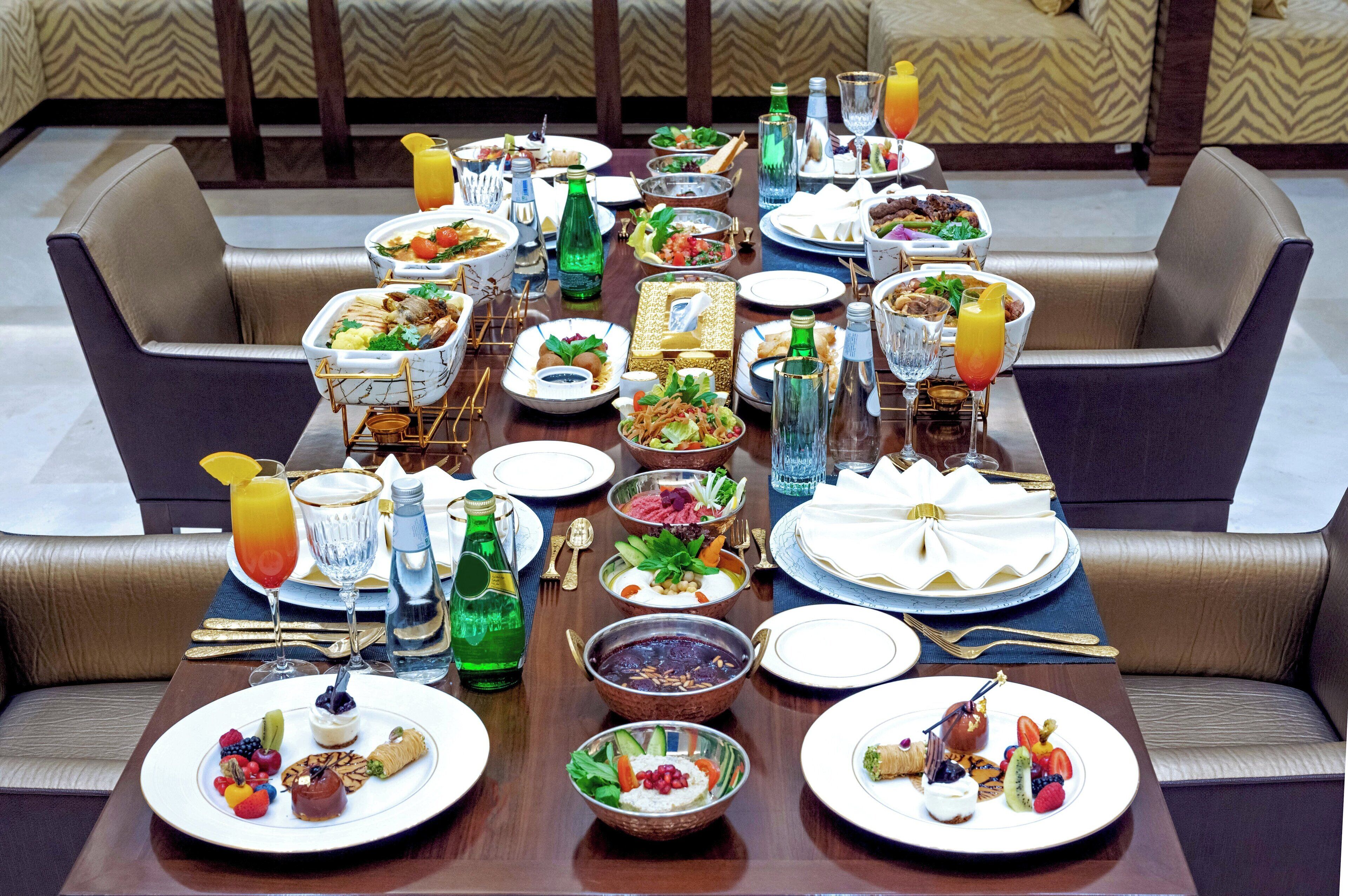 A dining table set with various dishes, drinks, and tableware, surrounded by upholstered chairs.