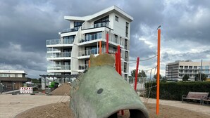Een speeltuin met een groot zandbak en klimconstructies, omringd door een verhard pad en een gebouw op de achtergrond.