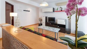 Una sala de estar moderna con una mesa de centro de madera, un televisor y una pequeña mesa de comedor con sillas. Hay una lámpara colgante y una planta en maceta sobre una estantería.