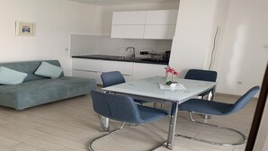A modern living room with a sofa, a dining table with chairs, a kitchen area, and a large window. - Residence Beni 6 with large heated pool (Medulin, Pula)