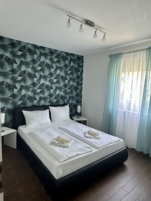 Een moderne hotelkamer met een hoofdeinde met een geometrisch patroon, een groot bed met witte lakens, twee nachtkastjes en een raam met dunne gordijnen.